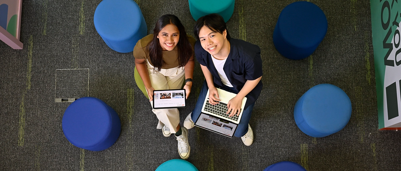 Top down view of 2 smiling students looking up from their computing devices.